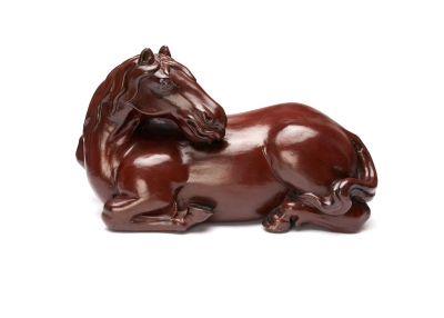 A dark brown shiny horse is lying on the ground with its legs curled underneath it and its head turned backwards. Its nose is resting on its back.